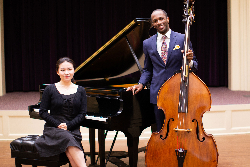 The Artists in Residence for 2024, Joshua and Yuhe. One is the woman, Yuhe, sitting down on a piano stool next to a piano while the other is a man, Joshua, holding a cello.